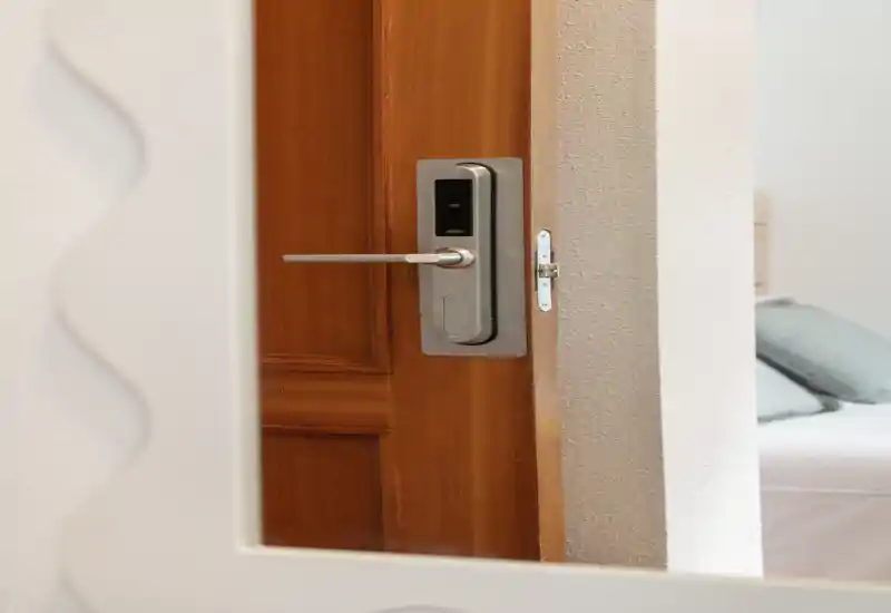 Vue partielle de la chambre à travers le miroir de la porte : un lit avec oreillers est visible sur le bord droit, la photo mettant l'accent sur la porte et la serrure.