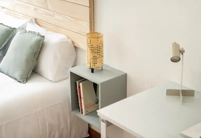 Rincón de dormitorio luminoso y minimalista con la cama bien hecha, cojines verdes, una lámpara de mimbre y una pequeña mesita con libros.