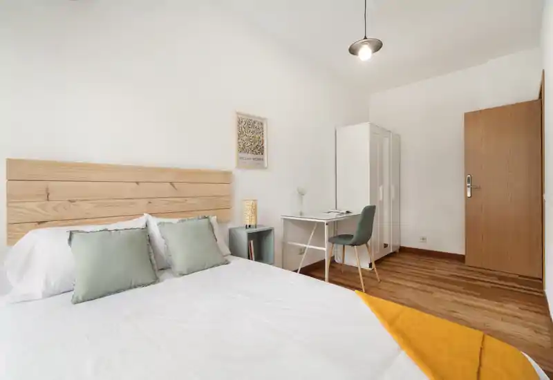 Dormitorio luminoso y minimalista con cabecero de madera, ropa de cama blanca y cojines verde suave. Incluye un escritorio y armario, ideal para trabajar y descansar.