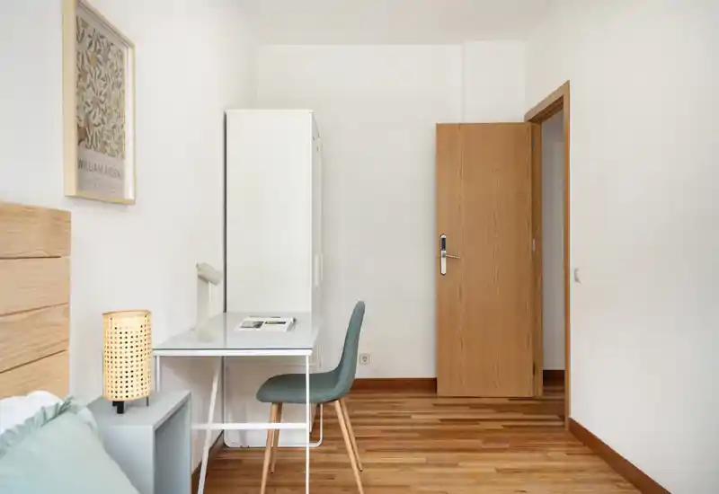 Coin chambre compact avec petit bureau, chaise et lampe de chevet. Décor neutre et sol en bois pour un espace calme et fonctionnel, adapté à une personne ou de courts séjours.
