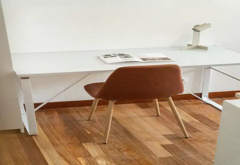 Espace de travail minimaliste avec un bureau blanc en verre et une chaise rembourrée marron sur un sol en bois chaleureux. Une lampe de bureau et un magazine ouvert apportent une touche fonctionnelle et soignée, parfait pour le travail concentré.