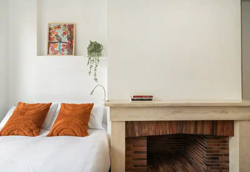 Camera da letto luminosa e minimal con letto ben rifatto, cuscini arancioni testurizzati e una pianta decorativa accanto al camino in pietra.