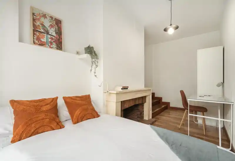 Camera da letto luminosa e ben arredata con letto bianco, cuscini arancioni caldi e un camino decorativo. È presente un piccolo angolo studio e un armadio, ideale per riposo e lavoro leggero.