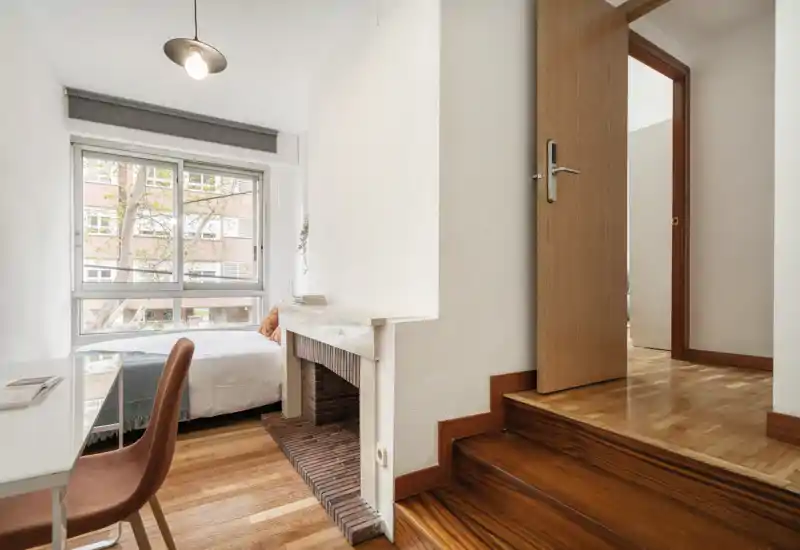 Camera da letto luminosa e compatta con ampia finestra, letto singolo con biancheria bianca, una piccola scrivania e pavimenti in legno caldo — accogliente e ben curata.