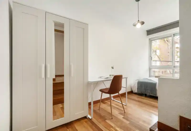 Chambre compacte et lumineuse avec un lit simple près de la fenêtre, un bureau minimaliste et une armoire à portes miroir. Parquet chaleureux et décoration neutre pour un espace fonctionnel et apaisant, idéal pour un étudiant ou une personne seule.