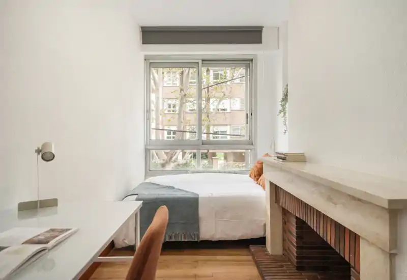 Camera da letto luminosa e minimal, con letto matrimoniale rivolto verso la finestra, biancheria morbida e dettagli caldi. Luce naturale abbondante e una scrivania semplice sul lato per lavorare o leggere.