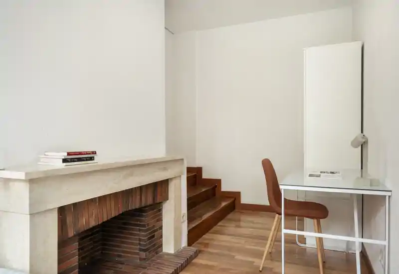 Espace de vie lumineux et minimaliste avec une cheminée classique, un sol en bois et un petit bureau contre le mur — un coin compact et fonctionnel pour travailler.