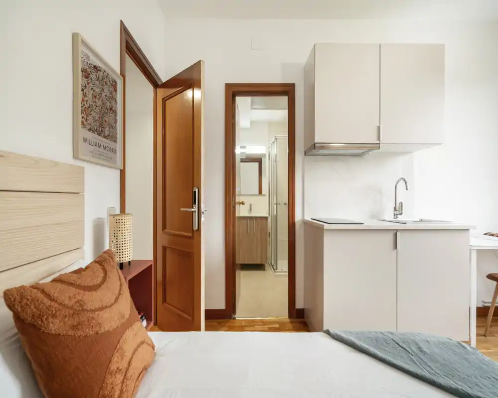Camera da letto luminosa e accogliente con letto ben visibile, tessuti caldi e angolo cucina compatto a destra; stile pulito e moderno, ideale per affitti brevi.