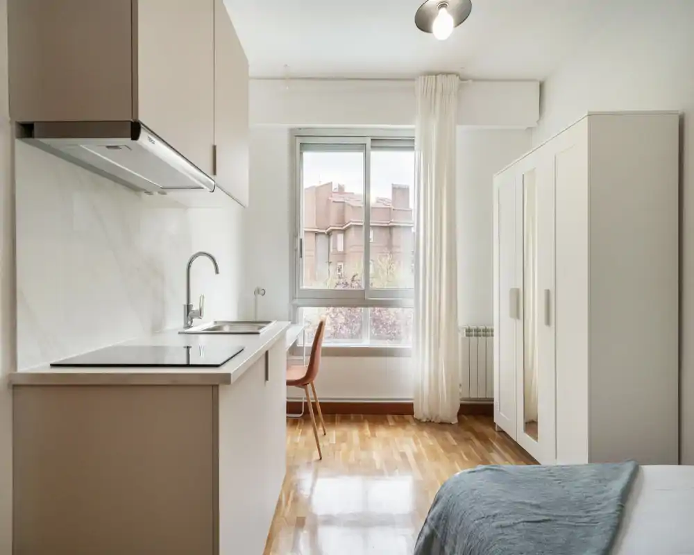 Camera da letto moderna e luminosa in stile monolocale con angolo cottura compatto, ampia finestra e armadio. L'ambiente è pulito e arioso con pavimento in legno e arredi minimal.