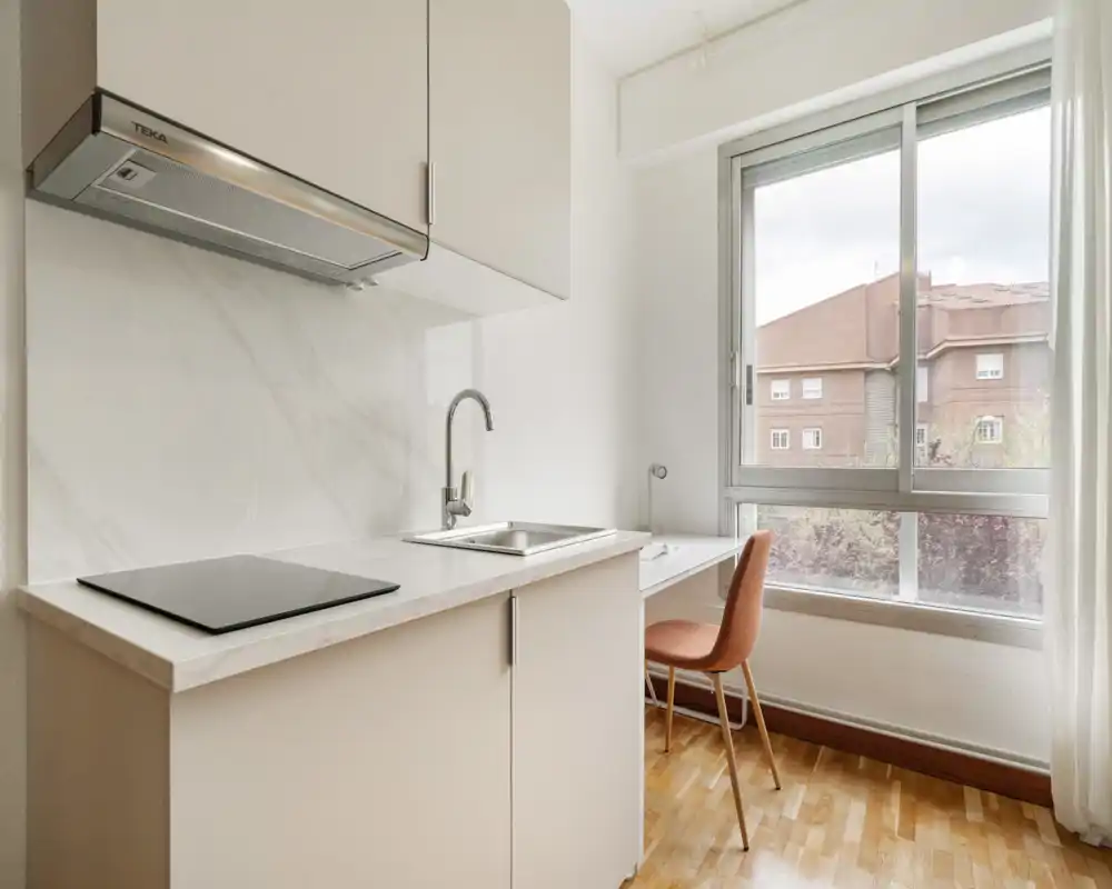 Kitchenette compatta e moderna con piano a induzione, lavello in acciaio e ampia finestra che illumina naturalmente; piccolo scrittoio e sedia rendono l'ambiente funzionale.
