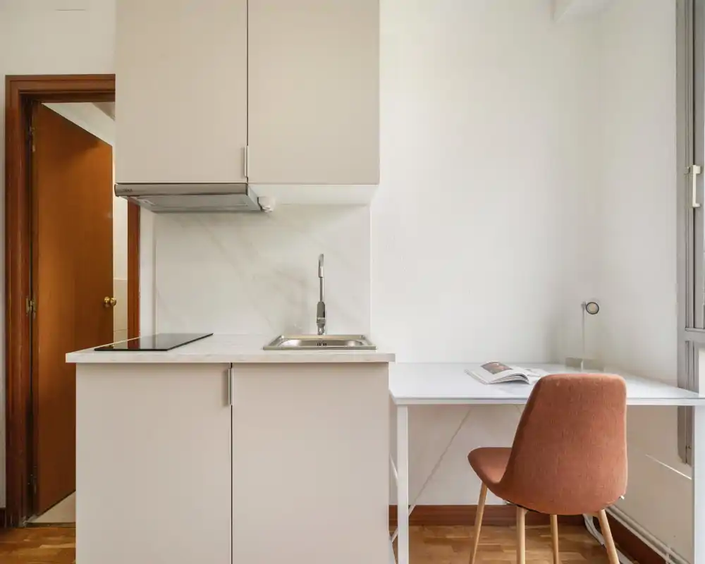 Angolo cottura moderno e compatto con lavello integrato, piano a induzione e una piccola scrivania accanto a una finestra luminosa — spazio pulito e funzionale ideale per un monolocale.