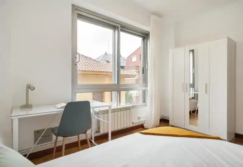 Dormitorio luminoso y ordenado con cama visible, escritorio junto a una gran ventana y armario independiente. La luz natural y los tonos neutros crean un ambiente tranquilo y funcional, perfecto para estancias relajantes.