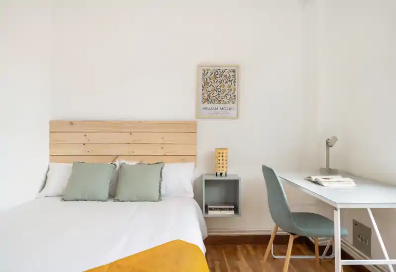 Dormitorio luminoso y minimalista con cabecero de madera, ropa de cama blanca, cojines verdes y un escritorio compacto — ideal para estancias relajantes y teletrabajo.