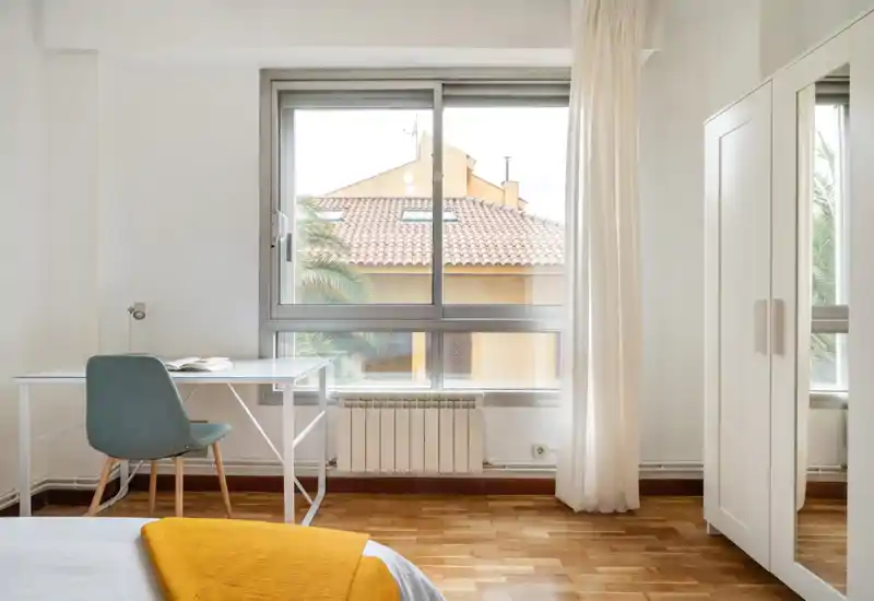 Chambre lumineuse et minimaliste avec grande fenêtre, bureau simple et armoire; parquet propre et un plaid moutarde apportant une touche chaleureuse.