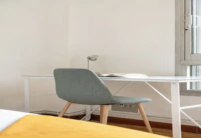 Coin de chambre lumineux et compact avec un bureau simple et une chaise moderne près d'une fenêtre ensoleillée ; un couvre-lit moutarde est partiellement visible au premier plan.