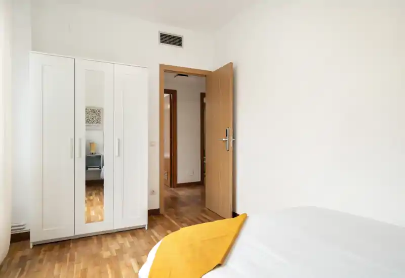 Chambre lumineuse et épurée avec coin de lit visible, armoire blanche avec miroir et parquet chaleureux—propre et accueillante pour locations courtes durées.