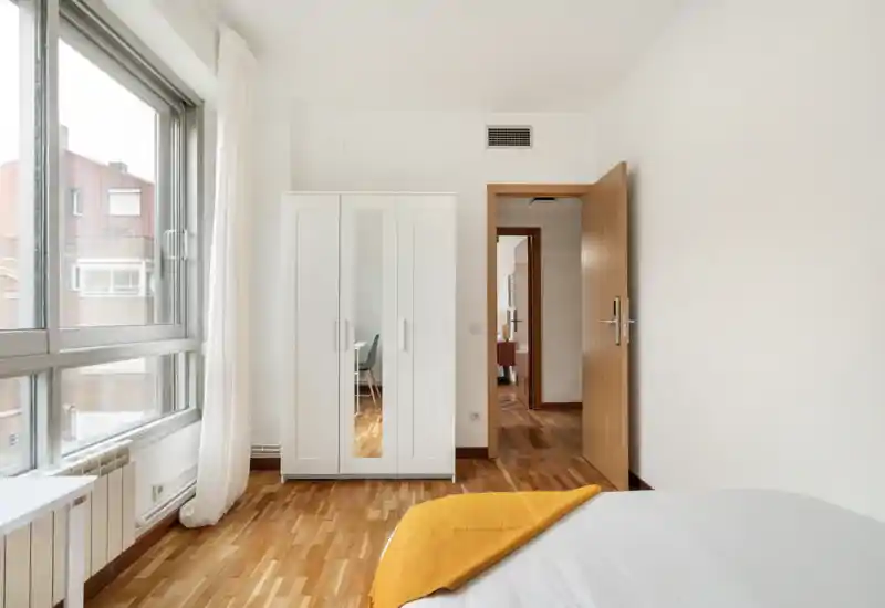 Dormitorio luminoso y minimalista con gran ventana, armario blanco con puerta espejo y cama con ropa blanca y manta mostaza — ideal para un anuncio moderno.