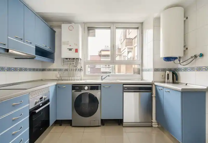 Cuisine lumineuse et soignée avec des façades bleues, des appareils en acier inoxydable et une grande fenêtre laissant entrer la lumière naturelle — pratique et bien organisée.