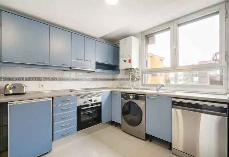 Cuisine moderne et lumineuse avec placards bleus, four intégré, lave-vaisselle et machine à laver sous le plan de travail; grande fenêtre apportant une belle lumière naturelle.