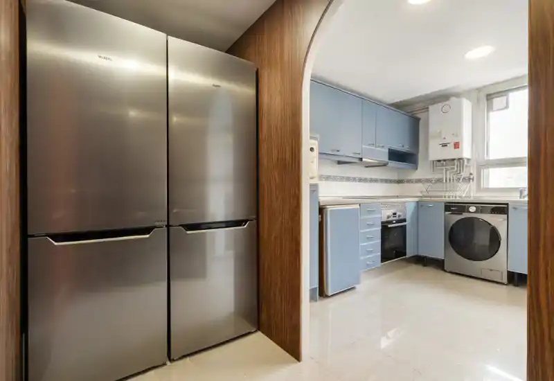 Cuisine moderne et compacte avec meubles bleus, four intégré, lave-linge et grand réfrigérateur inox; lumineuse, propre et pratique pour un usage quotidien.