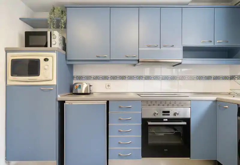 Cuisine compacte et lumineuse avec meubles bleus, four encastré, micro-ondes et petits appareils — propre et prête à l'utilisation quotidienne.