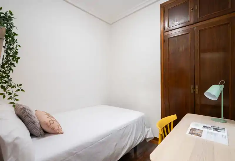 Dormitorio individual luminoso y ordenado con ropa de cama blanca, cojines decorativos, escritorio y armario empotrado de madera — espacio sencillo y acogedor.