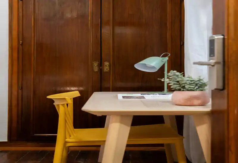 Pequeño rincón de estar/trabajo con mesa de madera, silla amarilla, lámpara de escritorio y planta en maceta frente a armarios de madera cálida.