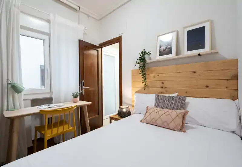 Dormitorio luminoso y acogedor con cabecero de madera, ropa de cama blanca y cojines decorativos. Un pequeño escritorio junto a la ventana y cuadros completan el espacio bien decorado.