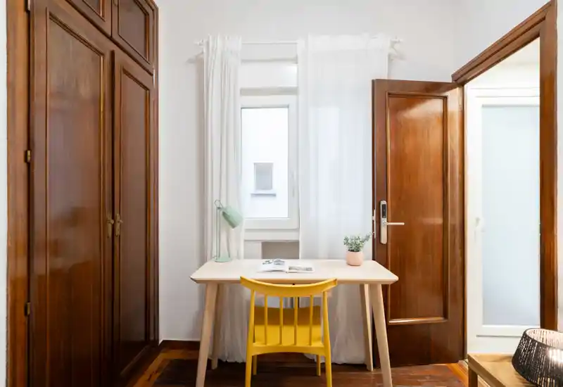 Habitación compacta con un pequeño escritorio frente a la ventana, puertas y armario de madera, con una sensación luminosa y aireada. La silla amarilla y la lámpara de mesa añaden un toque alegre al espacio minimalista.