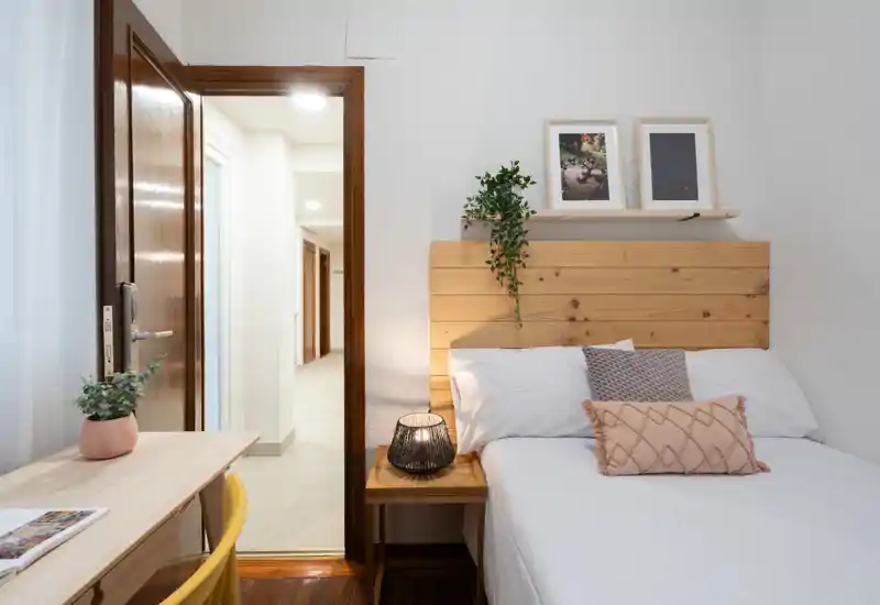 Dormitorio bien decorado con cabecero de madera, textiles acogedores y lámpara de noche; tonos claros y un pequeño escritorio que hacen el espacio funcional y acogedor.