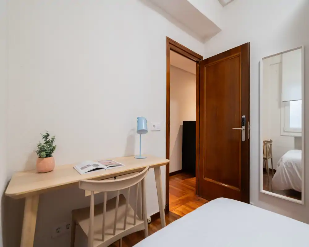 Chambre compacte et lumineuse avec un bureau simple, une chaise et le lit visible au premier plan. Décor neutre et un miroir pleine longueur près de la porte pour un espace propre et fonctionnel.