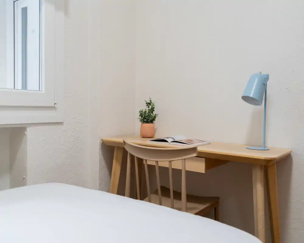 Coin de chambre lumineux et minimaliste avec le lit au premier plan et un bureau en bois équipé d'une lampe et d'une plante : un espace de travail propre et fonctionnel.
