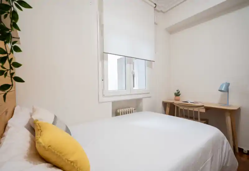 Dormitorio luminoso y compacto con cama doble bien hecha, cojines decorativos y un pequeño escritorio de madera junto a la ventana; estilo limpio y minimalista ideal para alquileres temporales.