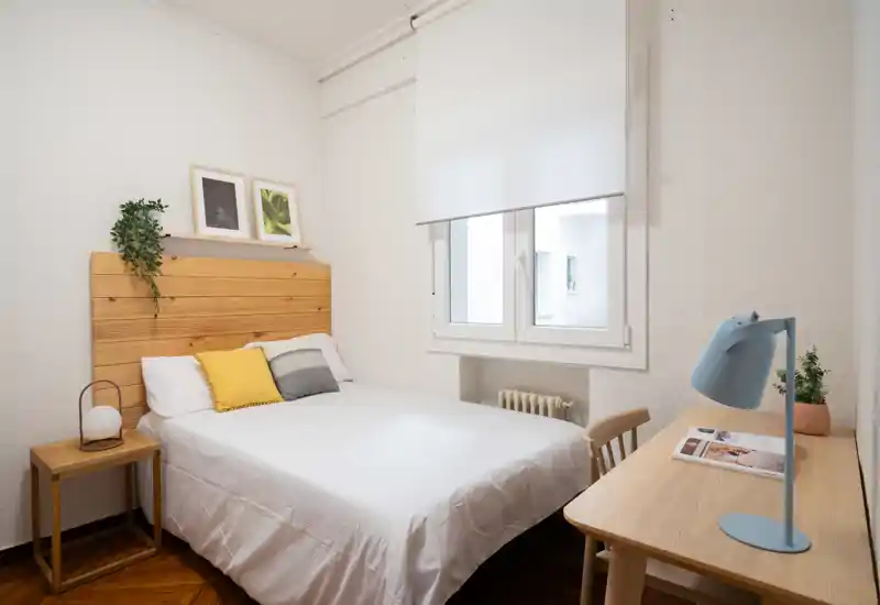 Dormitorio luminoso y ordenado con cama doble, cabecero de madera, mesita y escritorio junto a la ventana: acogedor y bien iluminado, perfecto para un anuncio.