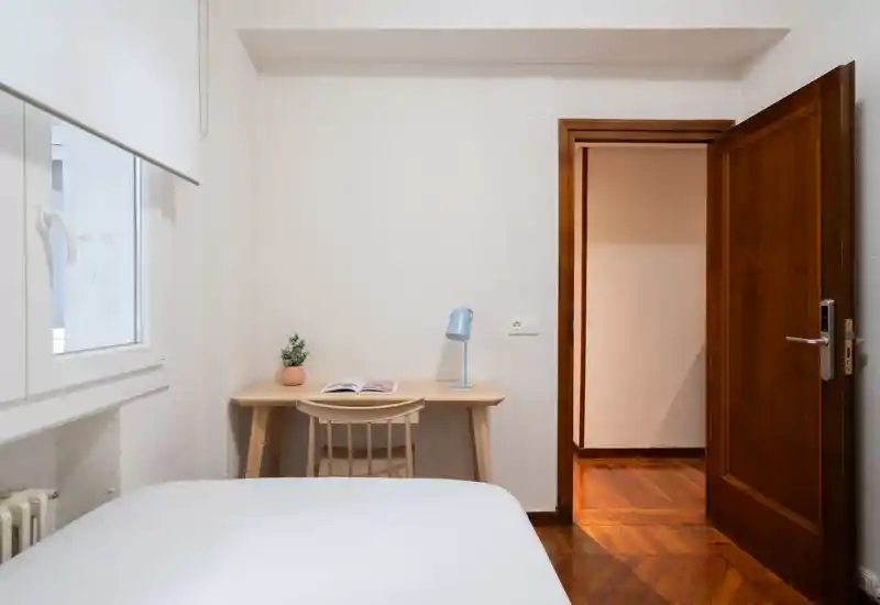 Dormitorio luminoso y de mobiliario sencillo con cama visible, pequeño escritorio de madera, lámpara y ventana que aporta luz natural — ideal para una estancia tranquila y minimalista.