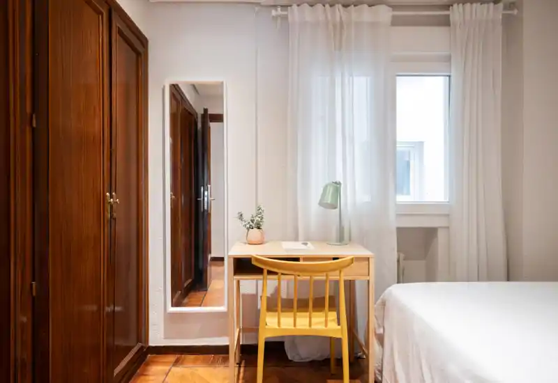 Bright, simply styled bedroom corner with a comfortable bed, wooden wardrobe, and a small desk by the window — ideal for a quiet work or rest spot.