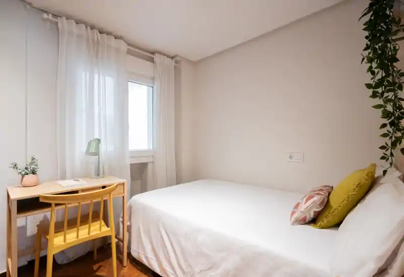 Bright, neatly staged bedroom with a comfortable double bed, two decorative cushions and a small wooden desk by the window letting in natural light.
