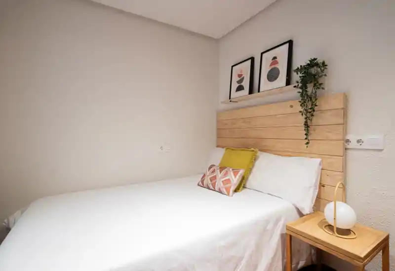 Cozy double bedroom with a wooden headboard, crisp white bedding and decorative cushions. Warm, minimalist styling and soft lighting make this room inviting for short stays.