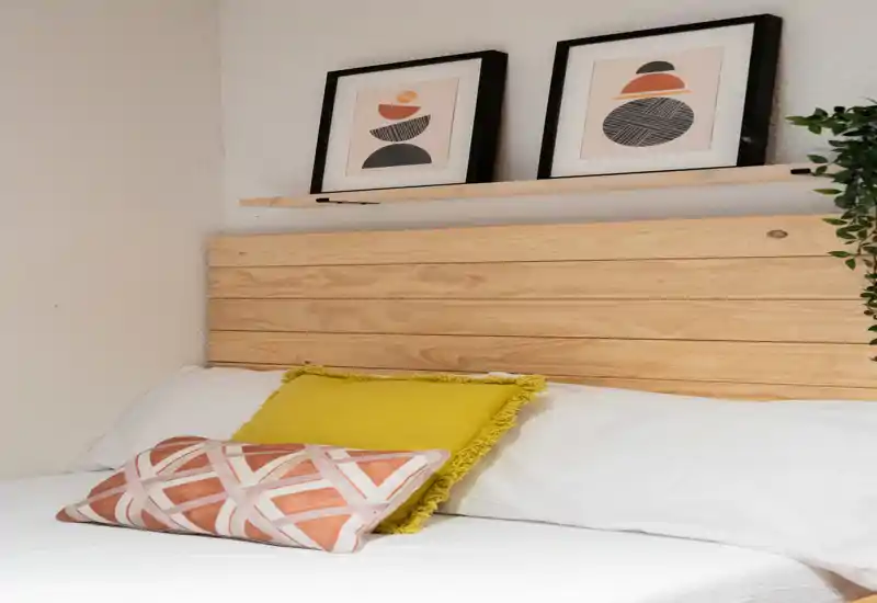 Bright, cozy bedroom corner with a wooden slatted headboard, decorative cushions and framed artwork — ideal for comfortable stays.