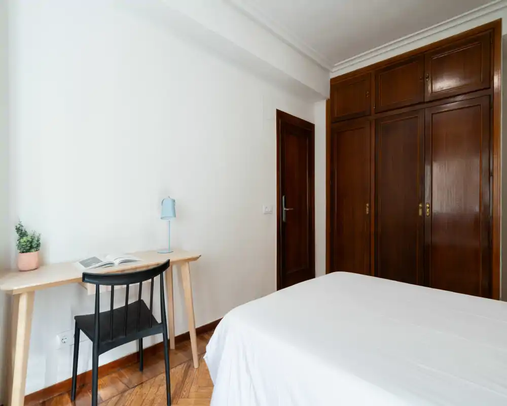 Camera da letto luminosa e ordinata con letto rifatto, armadio a muro in legno e una piccola scrivania con sedia e lampada — ideale per una singola o uno studente.