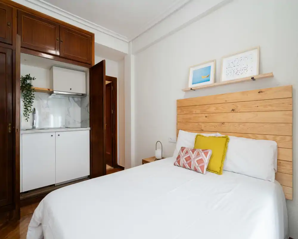 Camera luminosa e curata con testiera in legno e una piccola kitchenette integrata in un armadio rientrante — ideale per un monolocale in affitto.
