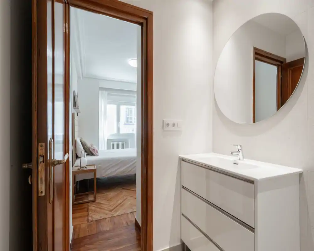 Camera da letto luminosa e moderna visibile attraverso una porta aperta, con letto confortevole, comodino e luce naturale dalla finestra. Pavimenti in legno caldo e palette neutra rendono l'ambiente accogliente.