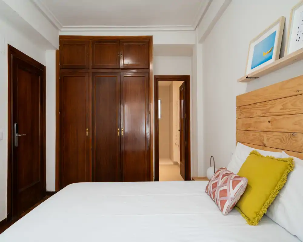 Camera da letto luminosa e ben arredata con testiera in legno, cuscini decorativi e armadio a muro in legno scuro; biancheria bianca e pareti neutre.