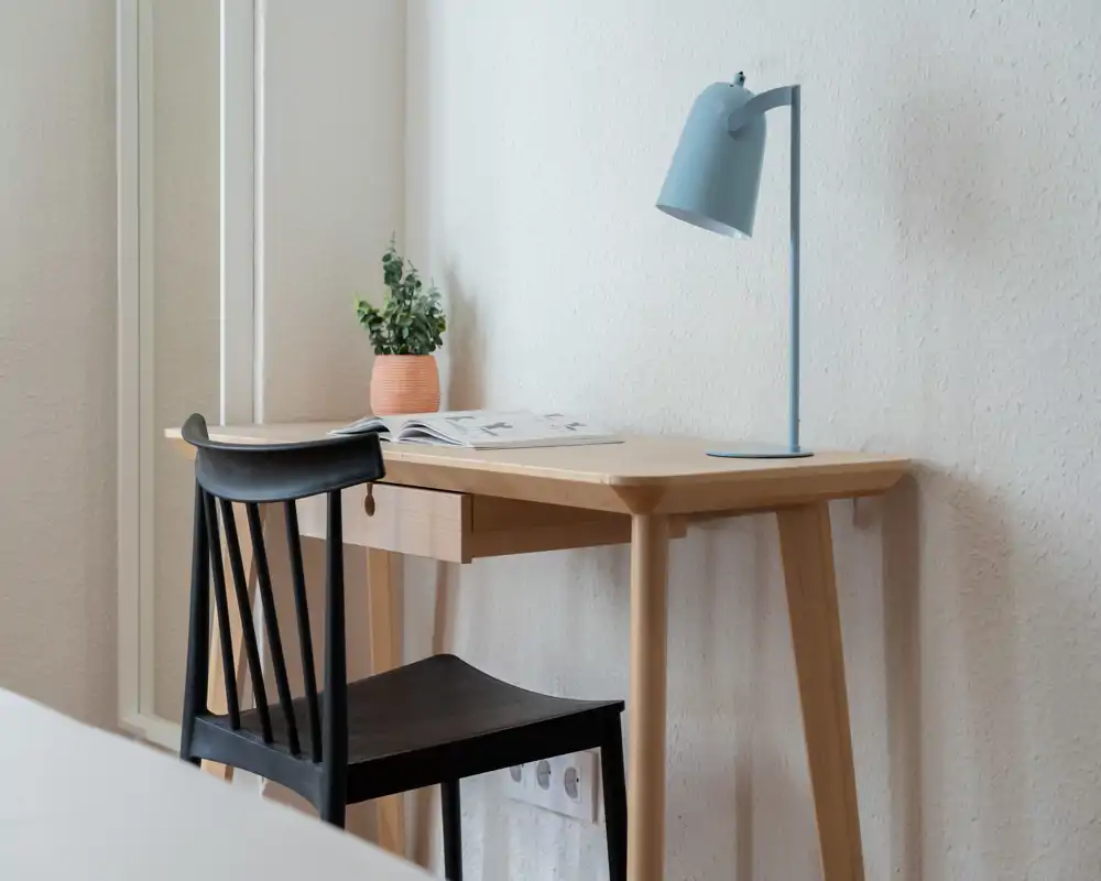 Angolo camera semplice con scrivania in legno, sedia nera, lampada da tavolo azzurra e piccolo vaso con pianta; una piccola porzione del letto è visibile sul bordo dell’inquadratura, creando un angolo lavoro ordinato e tranquillo.