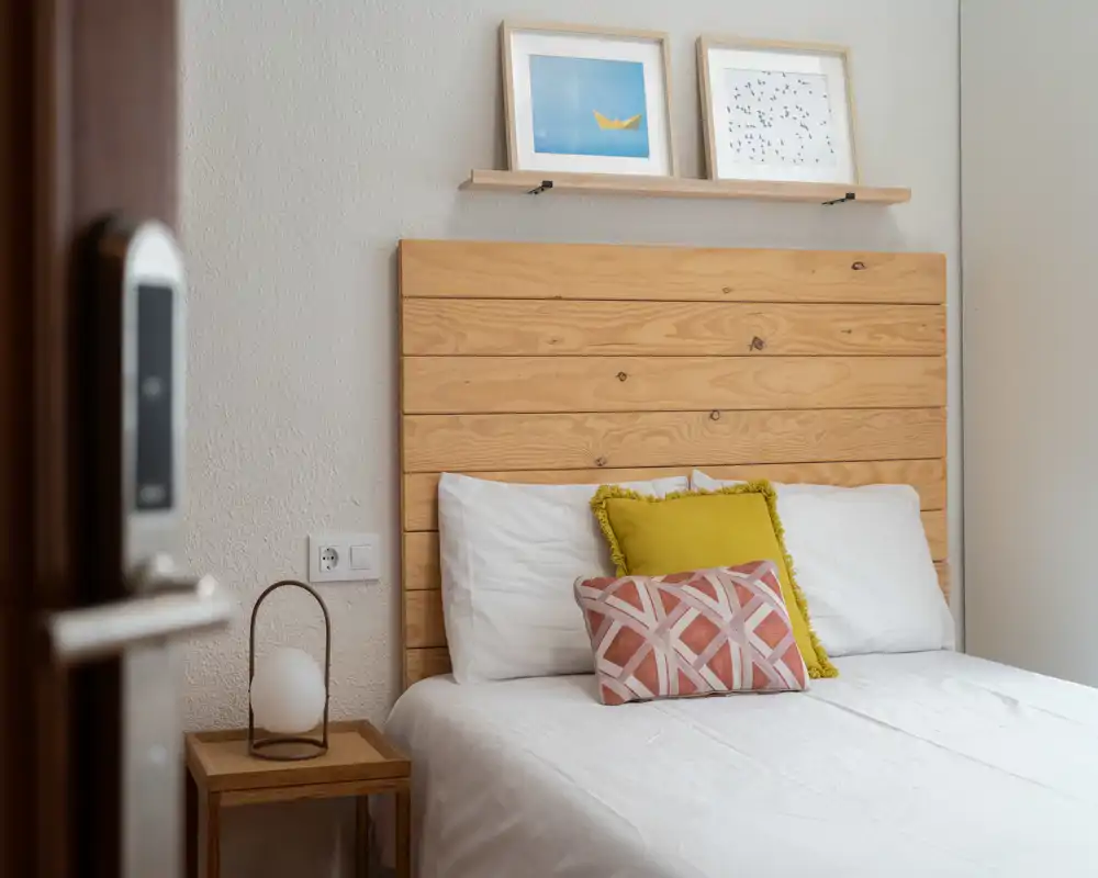 Camera da letto luminosa e accogliente con testata in doghe di legno, biancheria bianca e cuscini decorativi. Luce naturale morbida e arredo moderno essenziale creano un ambiente invitante.