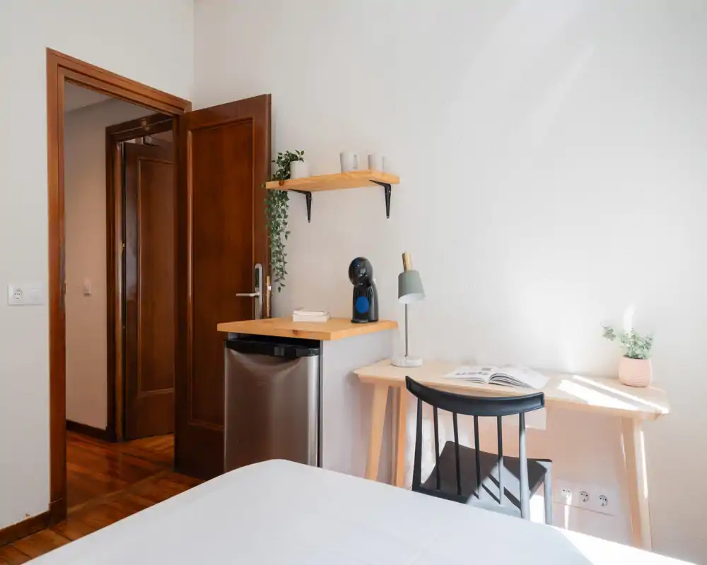 Rincón de dormitorio luminoso y ordenado con escritorio pequeño, silla, lámpara y mini nevera; la luz natural y el mobiliario sencillo crean un ambiente tranquilo junto a la cama.