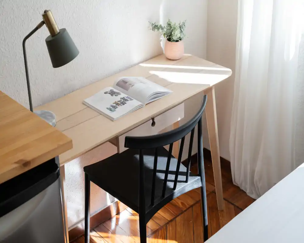Rincón de dormitorio luminoso y minimalista con mesa de trabajo de madera, silla, lámpara y una pequeña planta junto a cortinas traslúcidas; el borde de la cama se aprecia apenas a la derecha.