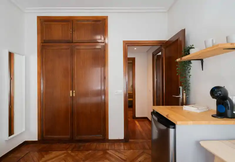 Pequeña zona de estar/entrada con armario empotrado de madera cálida, suelo de parquet y una encimera de cocina compacta con cafetera — espacio ordenado y funcional.