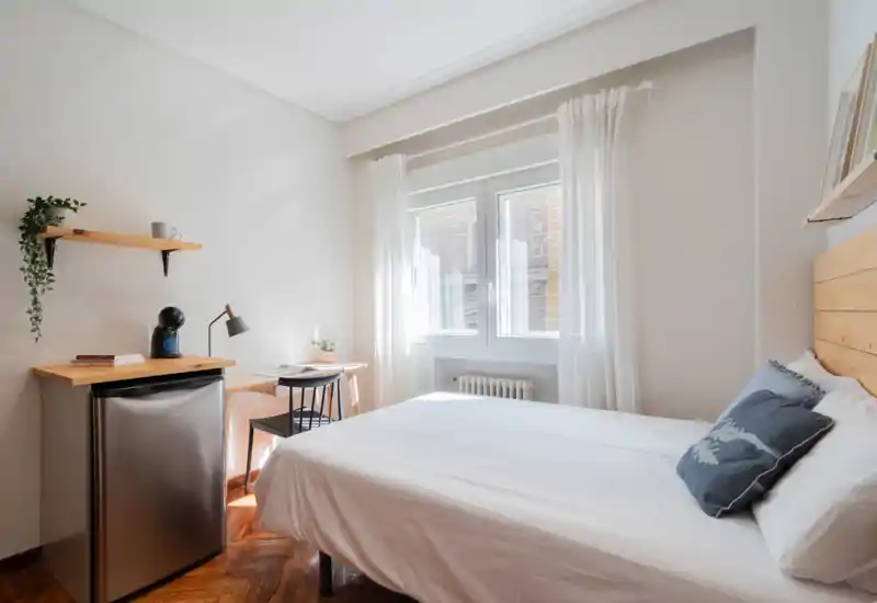 Dormitorio individual luminoso y bien equipado con cabecero de madera, ropa de cama blanca y un pequeño escritorio junto a la ventana — ideal para una estancia cómoda.