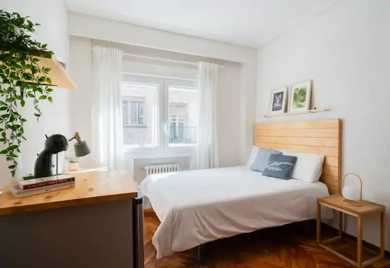 Dormitorio luminoso y minimalista con cabecero de madera, ropa de cama blanca y luz natural de una amplia ventana; decorado con una planta y detalles sencillos.